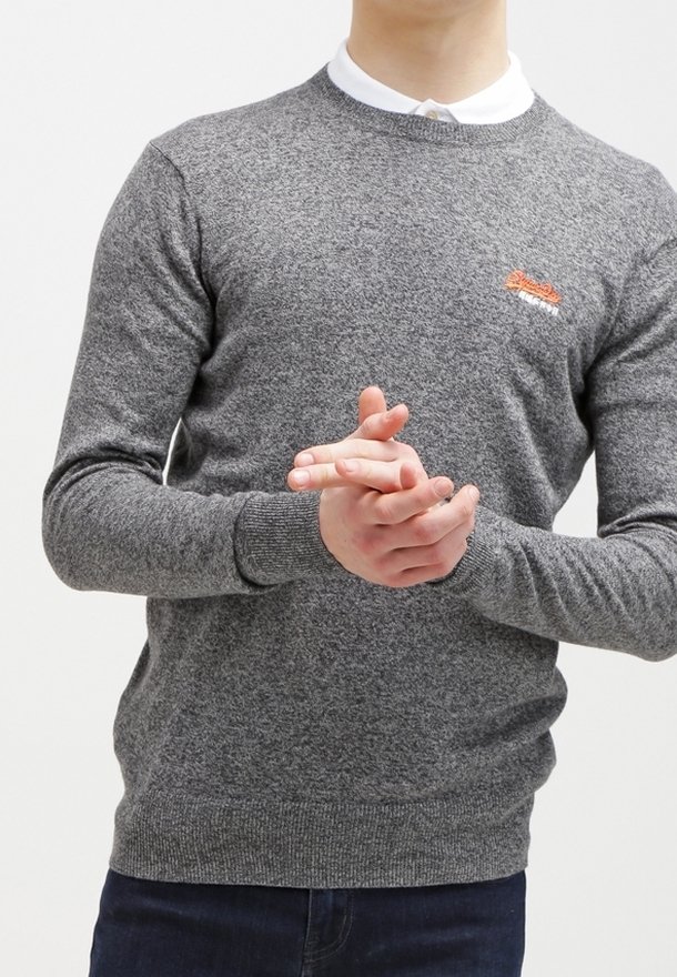 Personne portant un pull gris en maille par-dessus une chemise blanche à col, les mains jointes devant et un pantalon foncé visible.