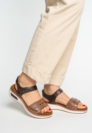 Feet wearing two-tone brown leather sandals with metal studs and beige corduroy pants against a plain light background.
