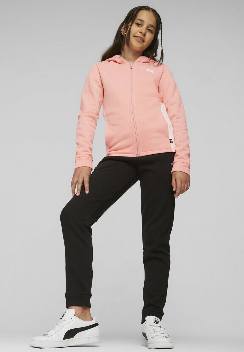 Felpa rosa con zip e accento bianco, pantaloni neri a costine e sneakers bianche con una striscia nera. La texture appare liscia e elastica.