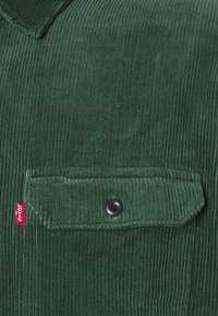 Tummanvihreä samettikangas, jossa on nappikiinnitys tasku ja pieni punainen kangastunnus taskun vasemmalla puolella.