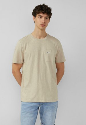 Młody mężczyzna o kręconych ciemnych włosach, ubrany w beżowy t-shirt z krótkim rękawem i jasnoniebieskie dżinsy, stojący na tle jednolitego jasnego tła.