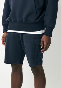 Marineblaue Cargo-Shorts aus Baumwolle mit einem lockeren Schnitt, ausgestattet mit Seitentaschen und einer flachen Vorderseite, kombiniert mit einem weißen T-Shirt darunter.