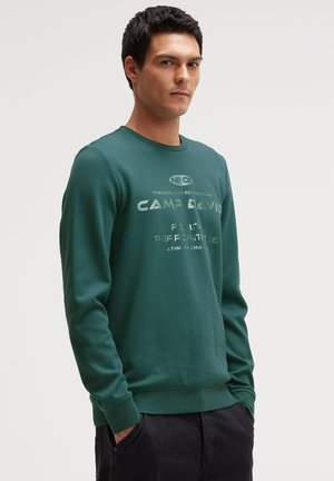 Man wearing a green long-sleeve sweatshirt with printed text, standing with hands in pockets against a plain background.