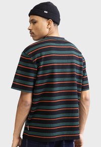 Camiseta de manga corta a rayas en verde oscuro, negro y naranja. Textura acanalada con un corte relajado y una pequeña etiqueta con el logo en el lado inferior.
