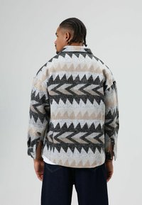 Grijze, beige en zwarte blouse met een geometrisch patroon, textuurstof, kraag en zijopeningen, gezien vanaf de achterkant.