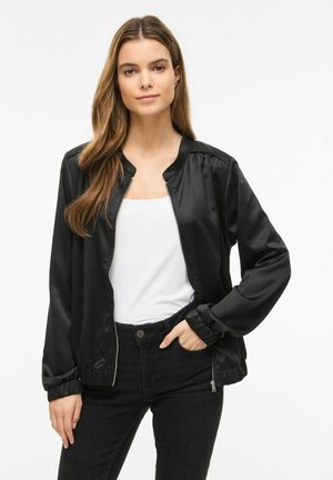 Kobieta ubrana w czarną satynową kurtkę bomberkę na białym topie i czarnych dżinsach, stojąca z jedną ręką w kieszeni na białym tle.