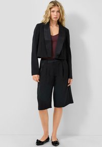 Blondīne, tērpusies melnā īsā žaketē, melnās ceļa garuma šortos, bordo kreklā un melnās lakotās kurpēs, stāv pie baltām sienām.