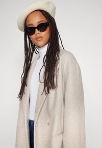 Hellbeige, übergroßer Wollmantel mit doppelreihiger Knopfleiste, kombiniert mit einem weißen Rollkragenpullover, schwarzen Katzenaugen-Sonnenbrillen und einer cremefarbenen Baskenmütze.