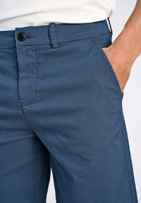 Pantaloni blu scuro con motivo, dalla texture liscia, dotati di chiusura con un pulsante e tasche laterali, mostrati in vita.