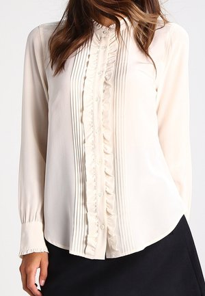 Vrouw draagt een crèmekleurige blouse met lange mouwen met verticale plooien en ruches langs de geknoopte voorkant, gecombineerd met een zwarte rok.