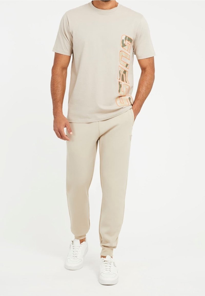 Beige Baumwoll-T-Shirt mit vertikalem "Guess"-Logo in Orange, kombiniert mit passenden beigen Jogginghosen und weißen Sneakers, im lässigen Schnitt.