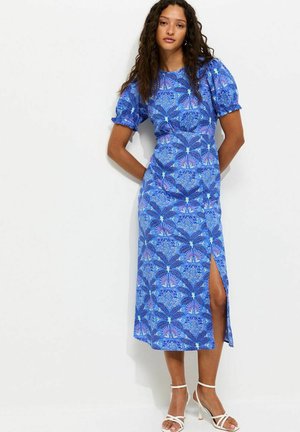 Kobieta o długich, kręconych włosach, ubrana w niebieską, wzorzystą sukienkę midi z bufkami i bocznym rozcięciem, stojąca na tle białego tła.