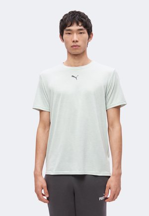 Jeune homme portant un t-shirt Puma gris clair et un pantalon de survêtement Puma foncé, debout sur un fond blanc uni.