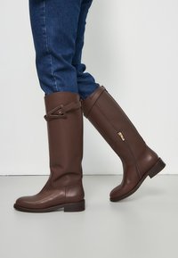 Brown knee-high leather boots with a smooth texture, side zipper, and decorative strap with a gold accent. Paired with blue denim.
