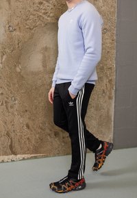 Světle modrá mikina, černé sportovní kalhoty s bílými pruhy a černo-oranžové sportovní boty. Hladká látka a moderní designové detaily.