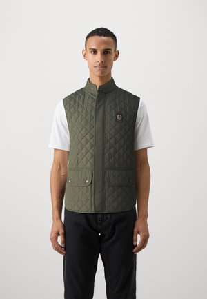 Gilet matelassé vert avec col haut, fermeture à pression sur le devant, deux poches avant et un petit logo sur la poitrine. Porté par-dessus une chemise blanche.
