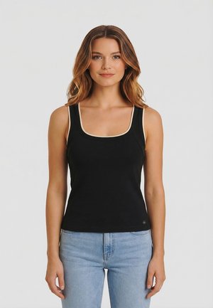 Mujer con cabello ondulado que lleva una blusa negra sin mangas con ribete beige y jeans azul claro, de pie contra un fondo claro y liso.