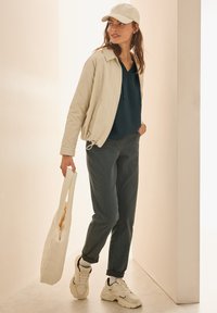 Beige gewatteerde jas, donkerblauwe hoodie met trekkoorden, grijze broek, witte totebag en stevige witte sneakers. Neutrale achtergrond.