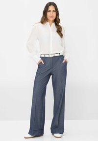 Witte blouse met lange mouwen en knoopsluiting, gecombineerd met marineblauwe, wijde broek met riem en zijzakken, tegen een effen achtergrond.