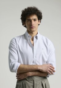 Camisa blanca a rayas con cuello abotonado, presenta finas rayas azules, mangas largas y un pequeño detalle del logo en el pecho.