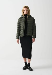Olivový prošívaný kabát s kapucí, prošívaný design, přední zip. Černé plisované midi šaty s lemem těsně nad kotníky a černé boty.
