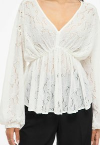 Biała koronkowa bluzka z dekoltem w serek, długimi bufkami i marszczoną talią. Przezroczysta tkanina ozdobiona wzorem kwiatowym, luźny i swobodny krój.