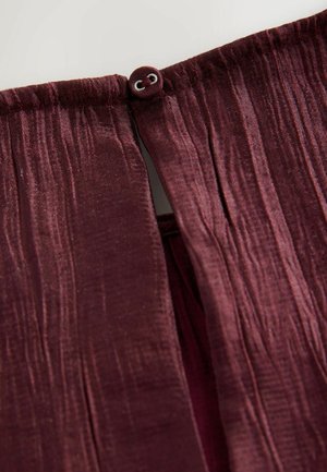 Burgundská skládací látka s mírně lesklou texturou a zapínáním na knoflík, která odhaluje vnitřní podšívku v podobné barvě.