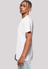 Junger Mann mit geflochtenem Haar, der ein schlichtes weißes T-Shirt und hellblaue Jeans trägt und im Profil vor einem einfarbigen Hintergrund steht.