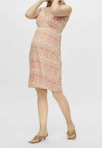 Persona embarazada que lleva un vestido sin mangas de largo hasta la rodilla y con un patrón, y sandalias de tacón bajo en color tostado, de pie frente a un fondo liso.