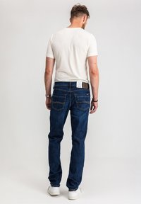 Ciemnoniebieskie dżinsy o prostym kroju, z firmową naszywką z tyłu, dwoma tylnymi kieszeniami i gładkim wykończeniem. Noszone z białym T-shirtem.