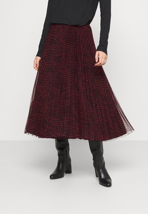 Kvinde iført en rød og sort plisseret midi-nederdel med ternet mønster, sort langærmet top og sorte knæhøje støvler med hæl.