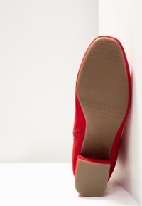Chaussure en daim rouge placée semelle vers le haut contre un mur blanc, affichant une semelle en caoutchouc beige avec "ANNA FIELD" embossé et un talon bloc.