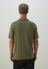 Khaki polo z krótkim rękawem z teksturowanej tkaniny, klasycznym kołnierzykiem i małymi rozcięciami po bokach u dołu, z czarnymi akcentami na kołnierzu.