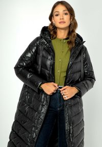 Schwarze Daunenjacke mit glänzender Textur, Kapuze und Reißverschlusstaschen. Sie hat ein langes, gestepptes Design und wird über einem grünen Pullover getragen.