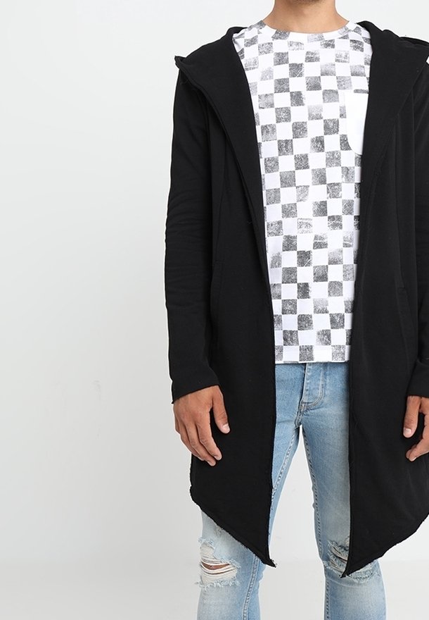 Cardigan nero lungo con cappuccio, con apertura frontale e tessuto texturizzato. Indossato sopra una t-shirt grafica bianca a quadri e jeans strappati.