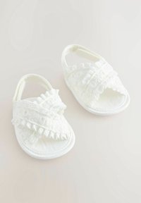 Bílé látkové sandály s křížovými pásky a volánkovými detaily. Plochá podrážka, otevřená špička a měkká textura pro pohodlí.