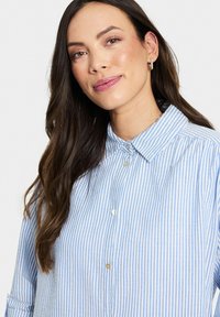 Mėlynos ir baltos dryžuotos marškiniai su apykakle, pagaminti iš lengvos medžiagos su subtiliu blizgesiu, papildyti perlų mygtukais.