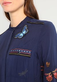 Mujer vistiendo una blusa azul marino con una mariposa azul bordada en el pecho y colorido bordado en la manga y la tapeta del bolsillo.