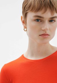 Camisa ajustada roja con escote redondo, llevada con pendientes de aro dorados. Textura suave; la modelo tiene el cabello corto y una expresión neutra.