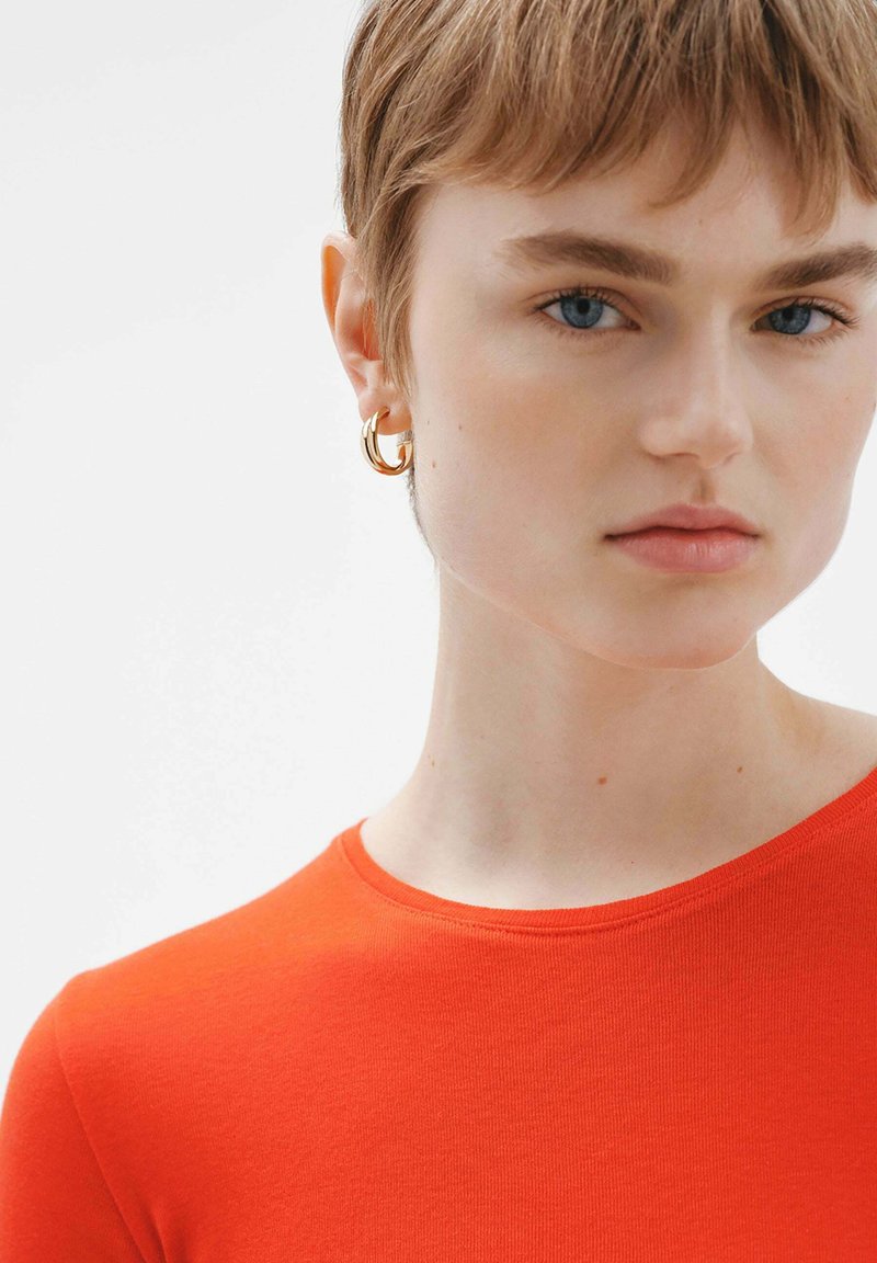 Camisa ajustada roja con escote redondo, llevada con pendientes de aro dorados. Textura suave; la modelo tiene el cabello corto y una expresión neutra.