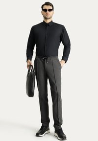 Maglietta nera a maniche lunghe, pantaloni grigi con coulisse, scarpe nere, che tiene una borsa nera. Occhiali da sole, texture lisce, vestibilità su misura.