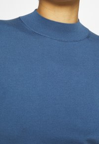 Niebieski, prążkowany sweter z wysokim kołnierzem, charakteryzujący się gładką teksturą i dopasowanym krojem. Zbliżenie na detale tkaniny i kołnierz.