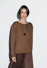 Eine Frau mit kurzem dunklem Haar, die einen braunen Langarmpullover, einen dunklen, plissierten Rock und eine schwarze Anhängerkette trägt, steht vor einer weißen Wand.