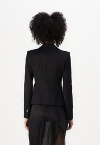 Černý přiléhavý blazer z hladké látky se strukturovanými rameny, zapínáním na jeden knoflík a decentním emblémem na rukávu.