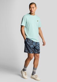 T-shirt di cotone azzurro chiaro con un piccolo logo giallo, abbinata a pantaloncini scuri a fantasia. Il modello indossa sneakers bianche e calzini a righe.