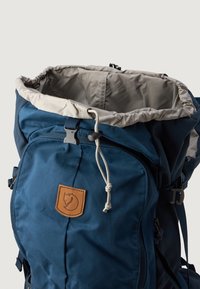 Σκούρο μπλε σακίδιο με ανοιχτότερο εσωτερικό, διαθέτει δερμάτινη ετικέτα λογότυπου, κλείσιμο με κορδόνι και πλαϊνές τσέπες για αποθήκευση.