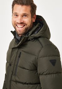 Dunkelgrüne Pufferjacke mit Kapuze, ausgestattet mit einem gesteppten Design, einer mit Reißverschluss versehenen Brusttasche und einem Markenlogo auf der Schulter. Strukturierten Stoff.