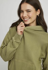 Sweat à capuche vert olive avec une poche avant et un cordon de serrage. Texture de tissu douce et poignets côtelés. Le mannequin tient la capuche près de son visage.
