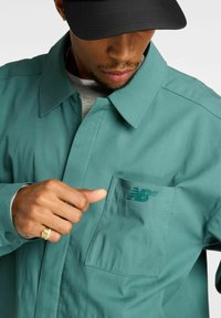 Giacca camicia turchese in tessuto liscio, con collo abbottonato, tasca frontale e logo in rilievo sul petto. Visibili gesti delle mani.