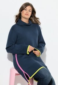 Maglione navy oversize con colletto a scollo, dettagli in neon giallo e rosa, polsini a costine e spacchi laterali, abbinato a jeans scuri.